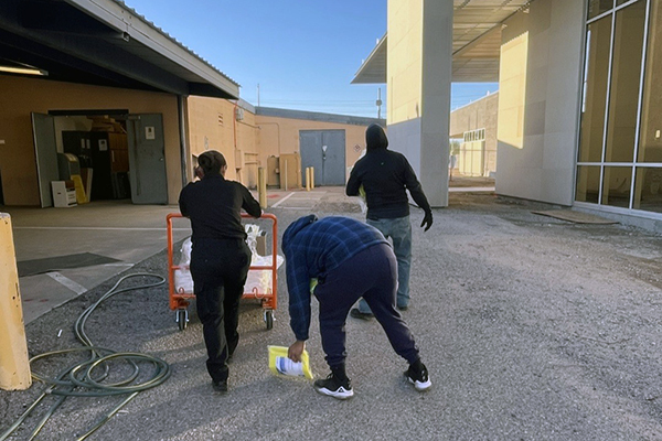 Three department members seen from behind, one pushing a dolly, one picking something up that dropped, head toward the new building.