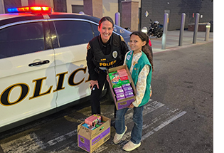 Smiling female officer and girl scout with boxes of cookies next to a patrol car.