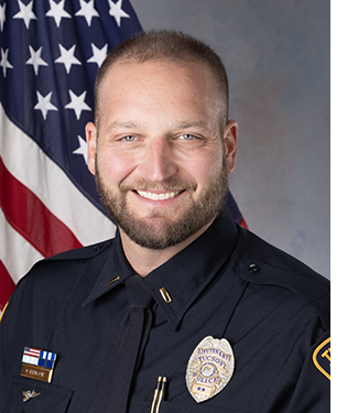 Department portrait of a male officer in uniform in front of an American flag.