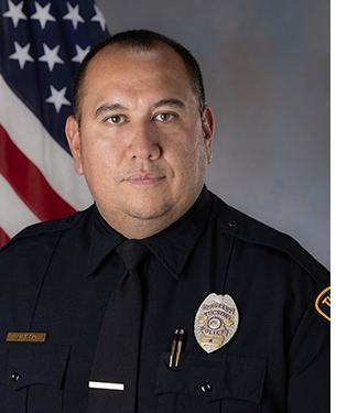 Department portrait of a male officer in uniform in front of an American flag.