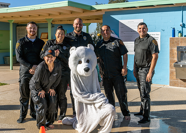 Officers in soaking wet uniforms pose for a group photo outside at a school. They are joined by a human in a polar bear suit.