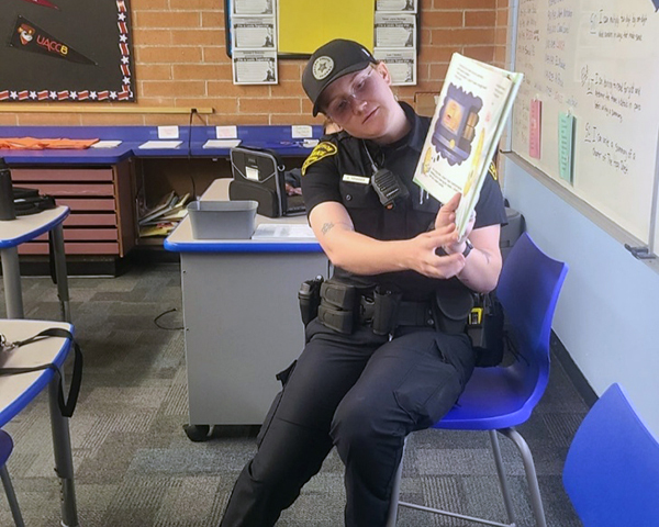 A female officer in uniform in a classroom reads to students from a children's book. She is showing them the illustrations.