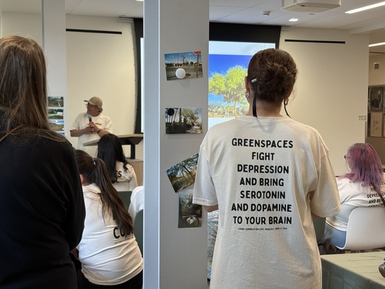Photo of A project participant addressing the crowd during the reception, while another wears a white graphic t-shirt. 