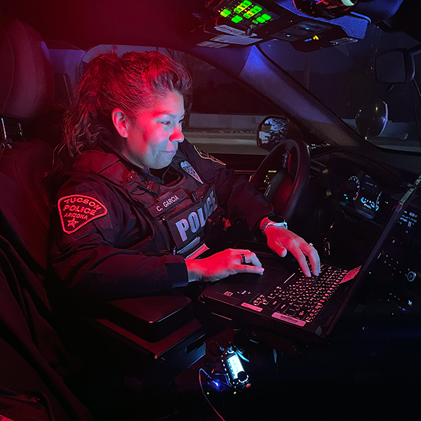 A female officer in uniform in a patrol car writes out a ticket on her department laptop. It is night and illuminated by the red/blue police lights.