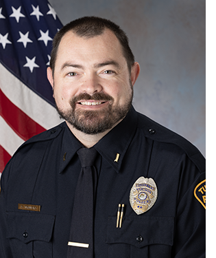Department portrait of a male officer in uniform in front of an American flag.