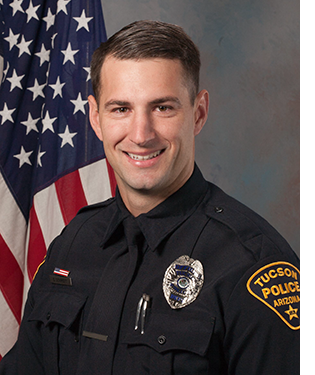 Department portrait of a male officer in uniform in front of an American flag.