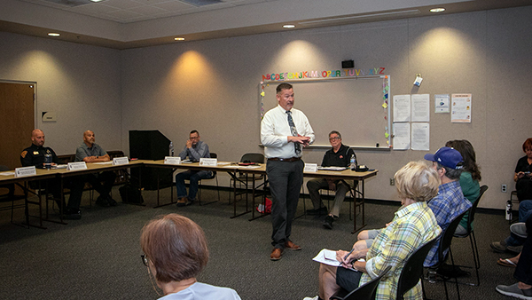 A man addresses a group in a public meeting space. Officers and community members are present.