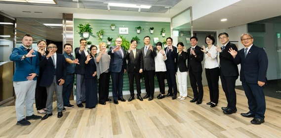 Seventeen men and women standing in an office setting and waving in front of a green wall with plants