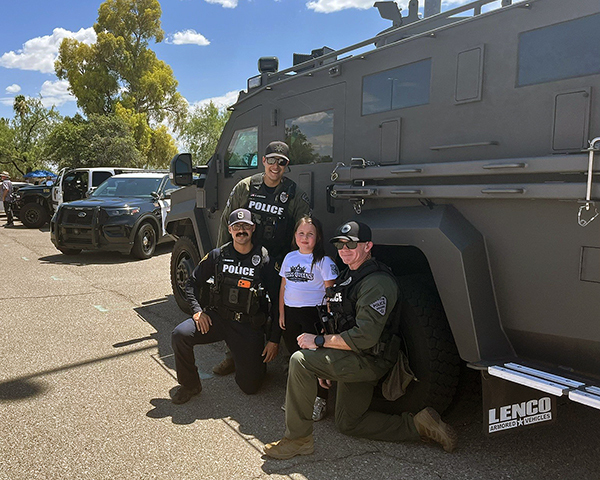 Police in uniform and one in SWAT uniform pose with a little girl in front of an armored vehicle.