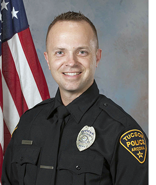 Department portrait of a male officer in uniform in front of an American flag.