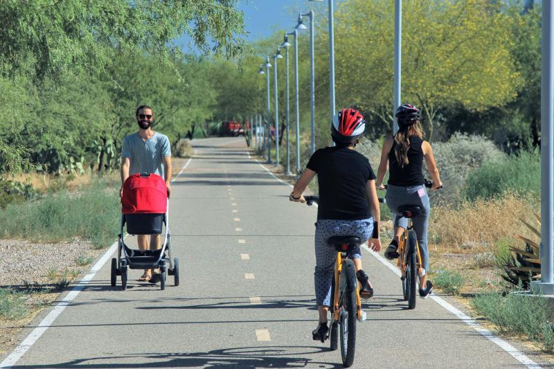Man pushing a red stroller walking and two women riding bicycles on a paved trail surrounded by greenery on a sunny day.