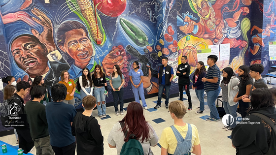 a group of 20-25 students stand in a circle with nametags on in front of a large, colorful mural