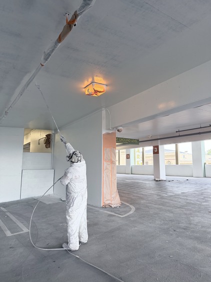 Photo of a worker in protective garb spraying white paint on the ceiling of a freshly painted parking garage interior