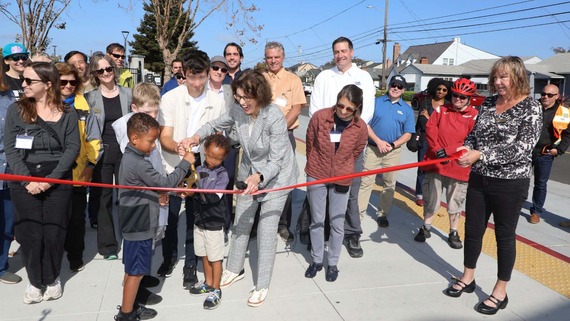 A group of people stand in front of a large red ribbon, while a few children hold ceremonial scissors and an adult holds a microphone