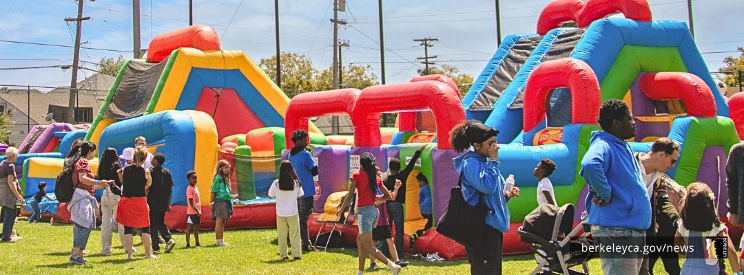 Children and families gather around colorful inflatable obstacle courses and bounce houses set up on a grassy field during a community event.