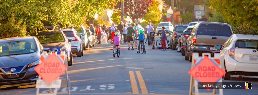 A street closed with orange barricades shows a group of neighbors meeting in the background for street block party.
