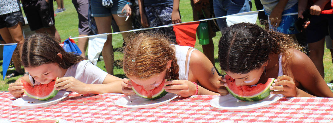 kids sitting at a picnic table eating watermelons without their hands