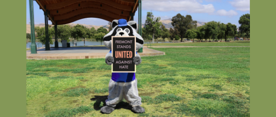 racoon mascot standing on lawn in front of outdoor pavilion at park and lake holding sign 