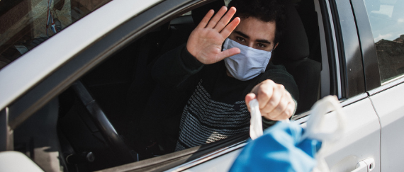 man in mask waiving from car to someone handing him a bag
