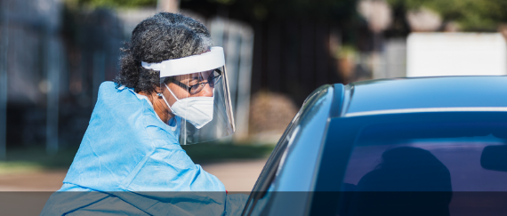 woman in gown administering COVID-19 test to person in vehicle