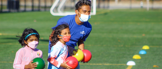 man with two kids in masks on sports field holding colorful balls 