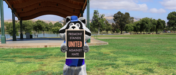RegeRec racoon holding 'Fremont Stands United Against Hate' sign in front of Central Park Performance Pavilion with lake and hills in background.