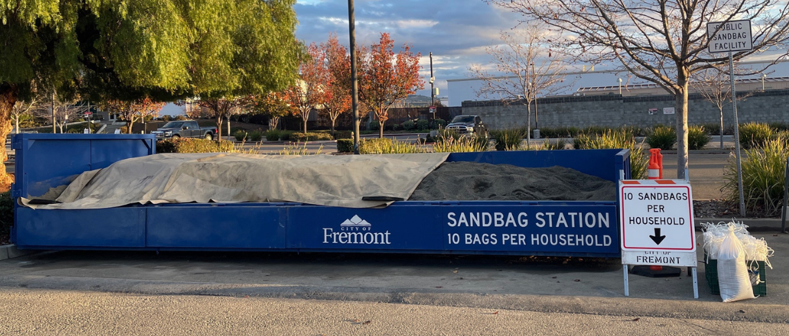 Mound of sand and bags for residents to make sandbags. Text: Sandbag Station. 10 Bags per Household.