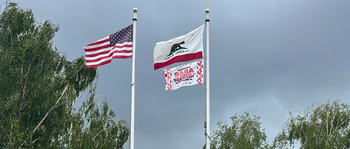 U.S. flag, State of California flag, and Arab American Heritage Month Flag on flagpole at City Hall with cloudy sky in background.  