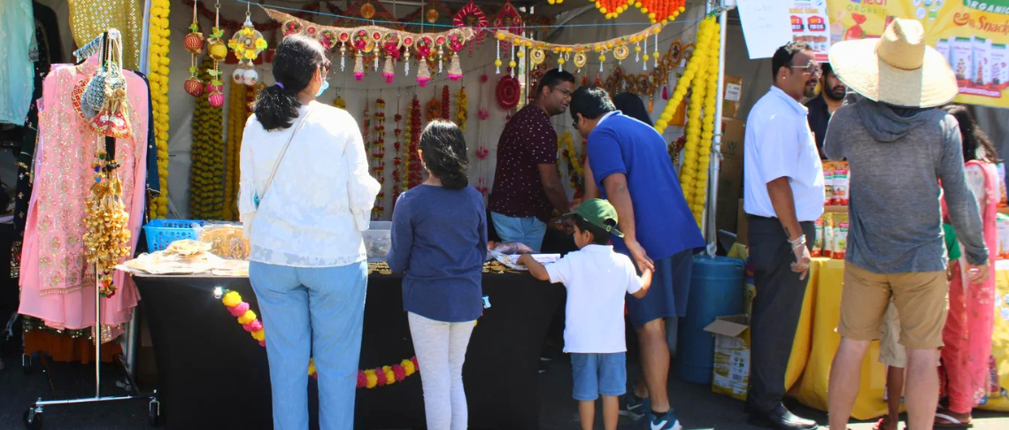 People browsing colorful handmade jewelry and decorations at an outdoor market stall on a sunny day.