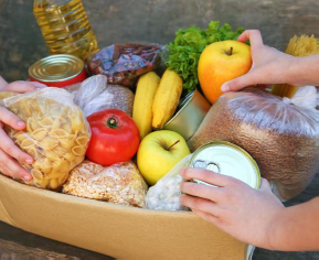 Two peoples' hands reaching into a basket if fresh food like apples, pomegranate, bread, dry pasta