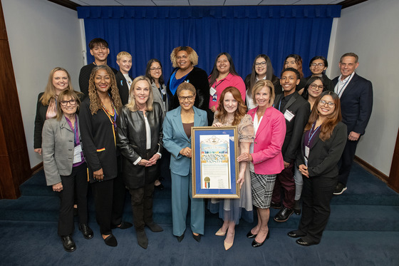 Supervisor Kathryn Barger stands with a large group of people with three women in the front holding a very large certificate in a frame