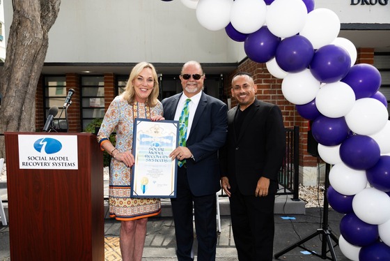 Supervisor Kathryn Barger stands with two men while holding a large certificate, beneath an archway of blue and white balloons 