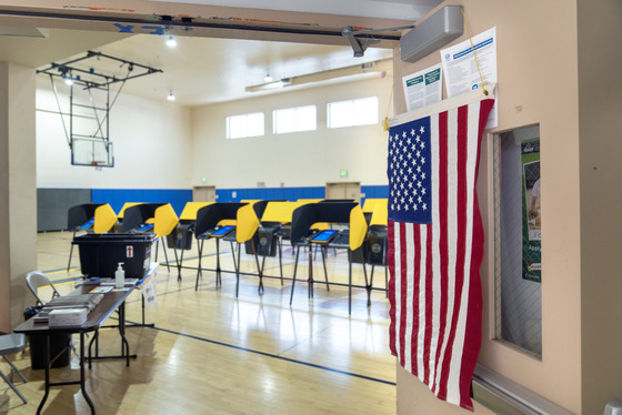 Front entrance to basketball court with flag United States flag hanging from door with a table across it, and yellow voting machines on the courts