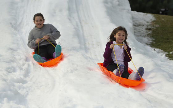 Two girls sliding on orange sleds down white snow at Charter Oak Park in Covina