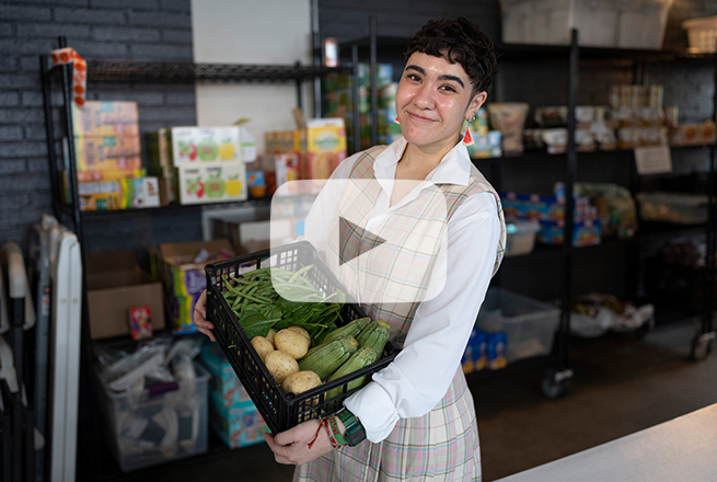 Person with short hair a white long sleeve shirt, and a checkered brown dress holding a basket with vegetables