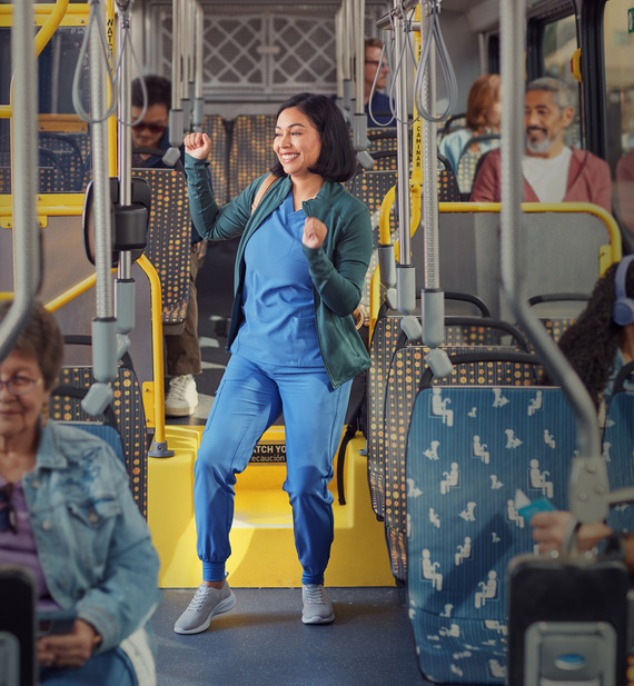 Woman wearing blue nursing uniform and green sweater is standing up on the Metro bus with people sitting around her