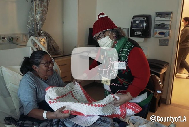 Woman with red beanie and red Christmas sweater give a hand-made quilt to a female patient at LA General Medical Center