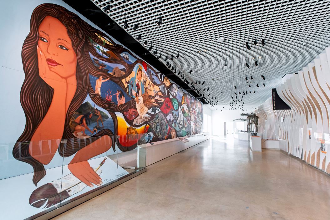 Mural of a woman and her long hair leads to other images on the mural, white ceiling, white wall across, and a dinosaur far out 