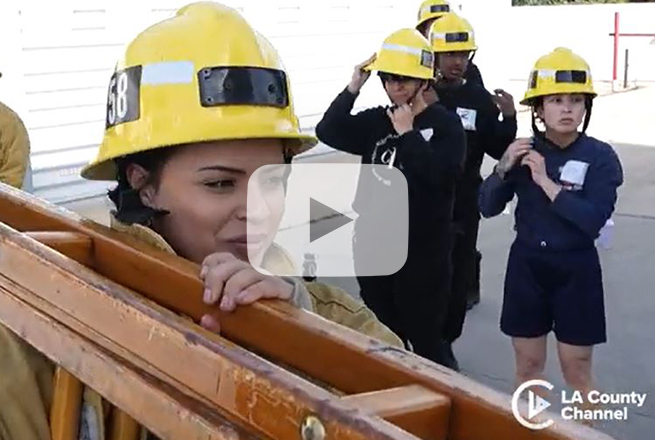 Female wearing yellow helmet, yellow jacket and holding a ladder on her shoulder while other females with helmets stand