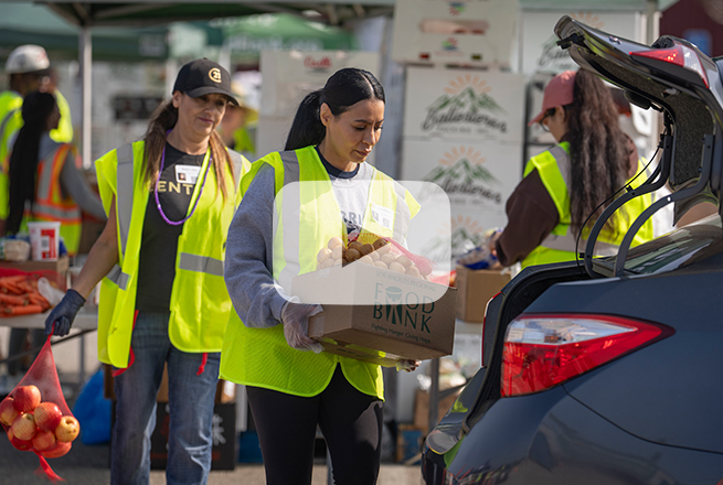 Woman carrying box of food towards the trunk of a car and another woman behind her carrying a bag of potatoes
