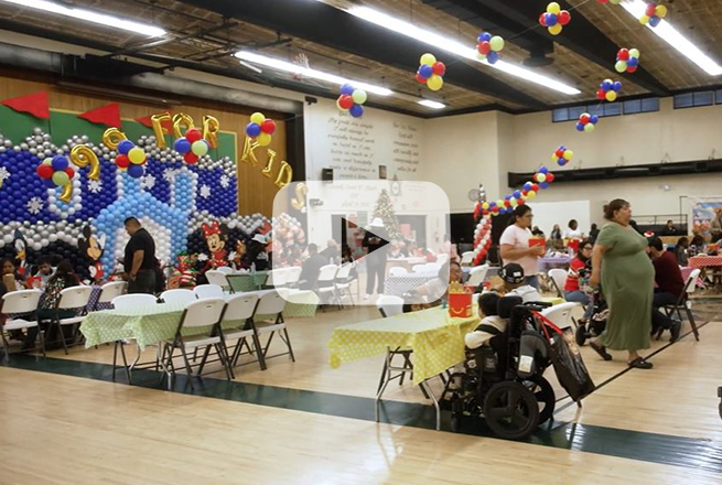 Families sitting at tables with colorful table cloths, balloons hanging from the ceiling and a Christmas tree in the corner of the room