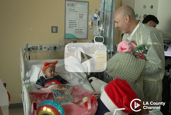 Girl laying in hospital bed looks up at man standing next to her holding hand made quilts
