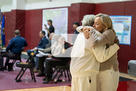 Two women hugging inside a gymnasium with people sitting on chairs in the background 