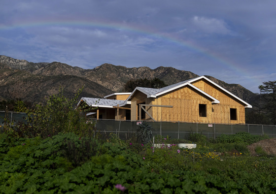 Wooden frame of house being built with green fence surrounding it, mountain and rainbow behind it