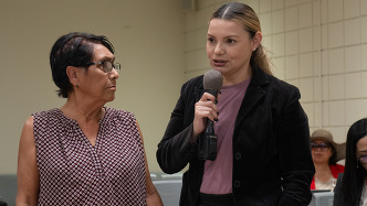 Two women at a public meeting, one speaking into a handheld microphone while the other stands beside her listening.
