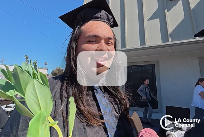 Man with long hair, wearing cap and gown, tie, and holding a plant