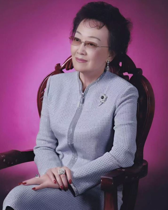 a woman in a gray suit jack seated in a chair, with a purple background