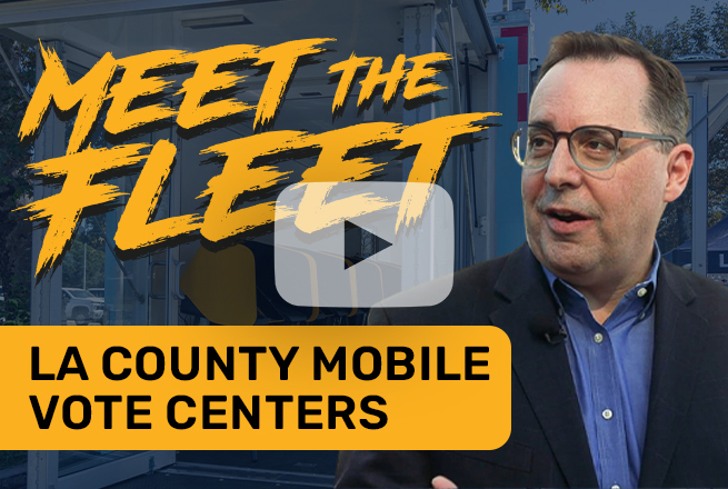 Man wearing glasses, dress shirt, suit jacket and in the background a mobile voting trailer
