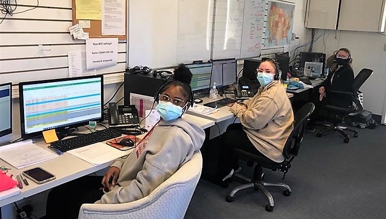 Three women wearing face coverings sit at computer work stations with headsets on at the call center.