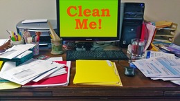 A computer monitor with the words "Clean Me" on a messy desk full of papers.